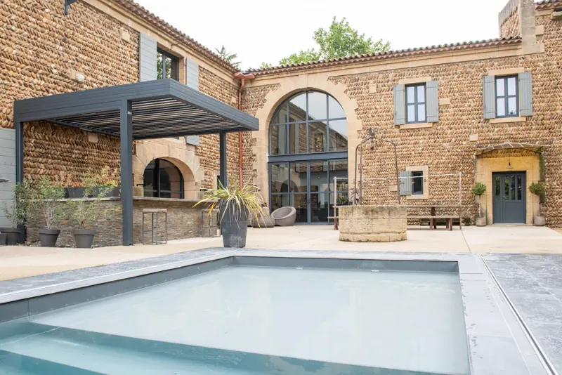 Pergola à lames orientables grises posée devant une bâtisse en pierre, au bord d’une piscine